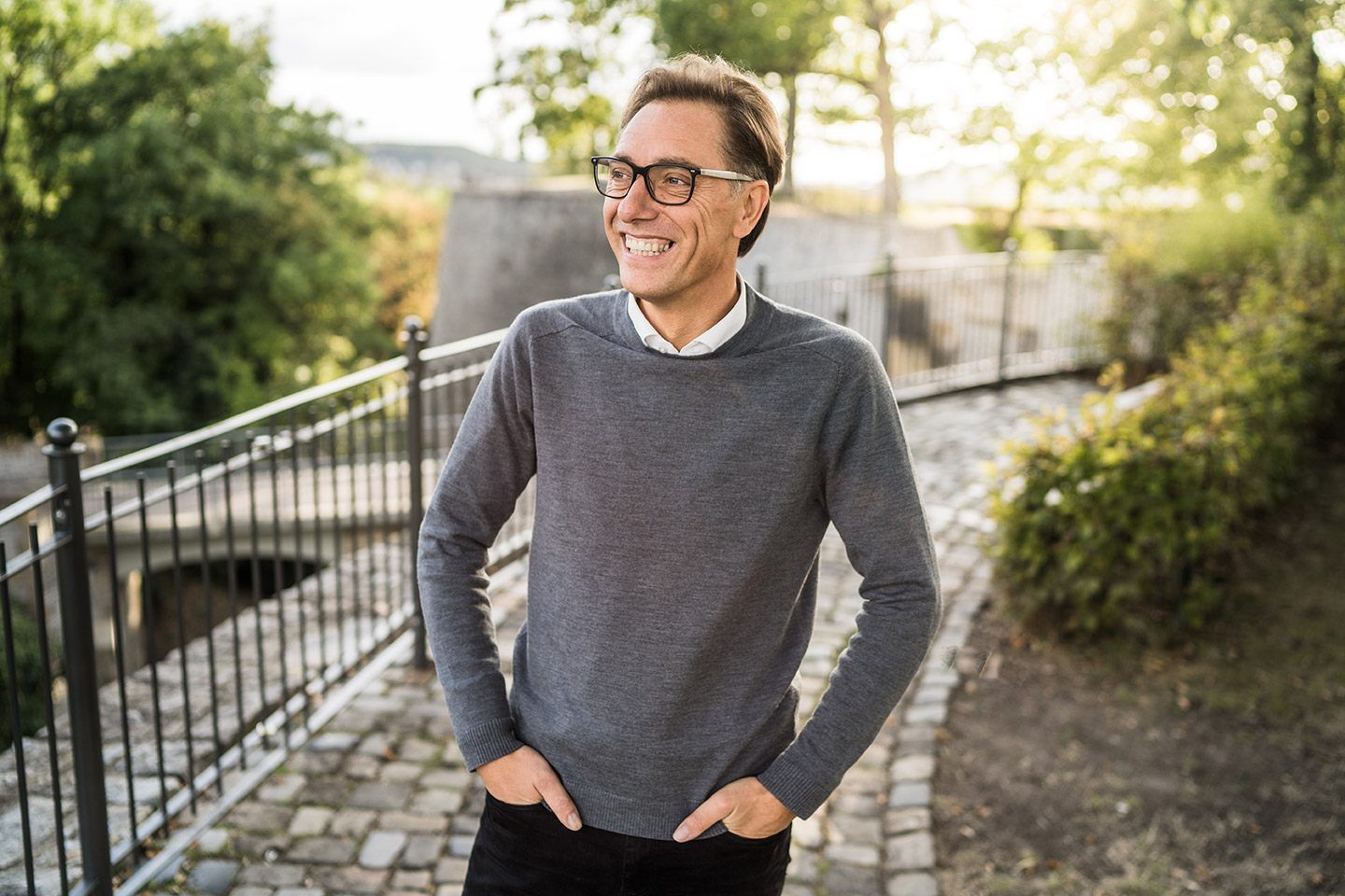 René Müller lacht an einem Geländer im Park, grauer Pullover, Brille